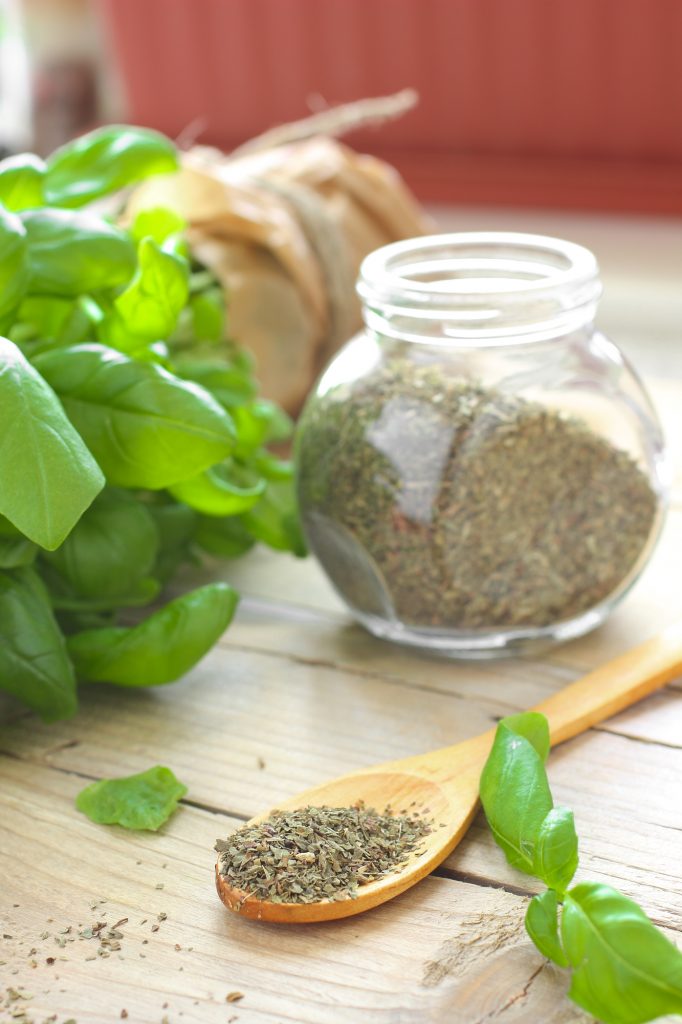 dried basil on a wooden spoon in front of a glass jar of basil and a basil plant