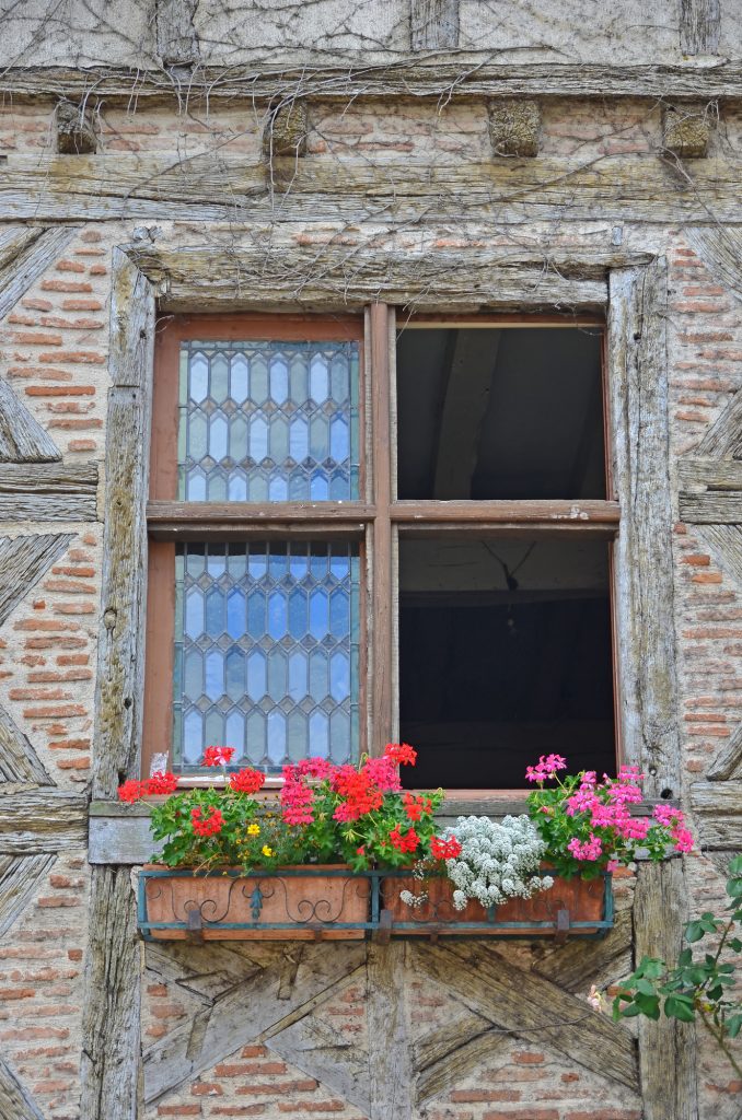 Window in traditional half timbered house decorated with a geranium filled window box, lead panel windows, and simple saltire and brick filled walls